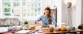 Woman using scissors sat at a table covered with boxes, jars, balls of wool and folders